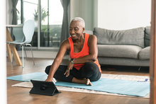 A beautiful senior woman takes an online yoga class. She is interacting with the teacher on her tablet and about to commence her class. She is wearing casual active wear and is taking the class in her lounge room. She is sitting on a blue yoga mat.