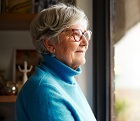 An elderly woman stands next to a window and looks outside.
View with copy space.